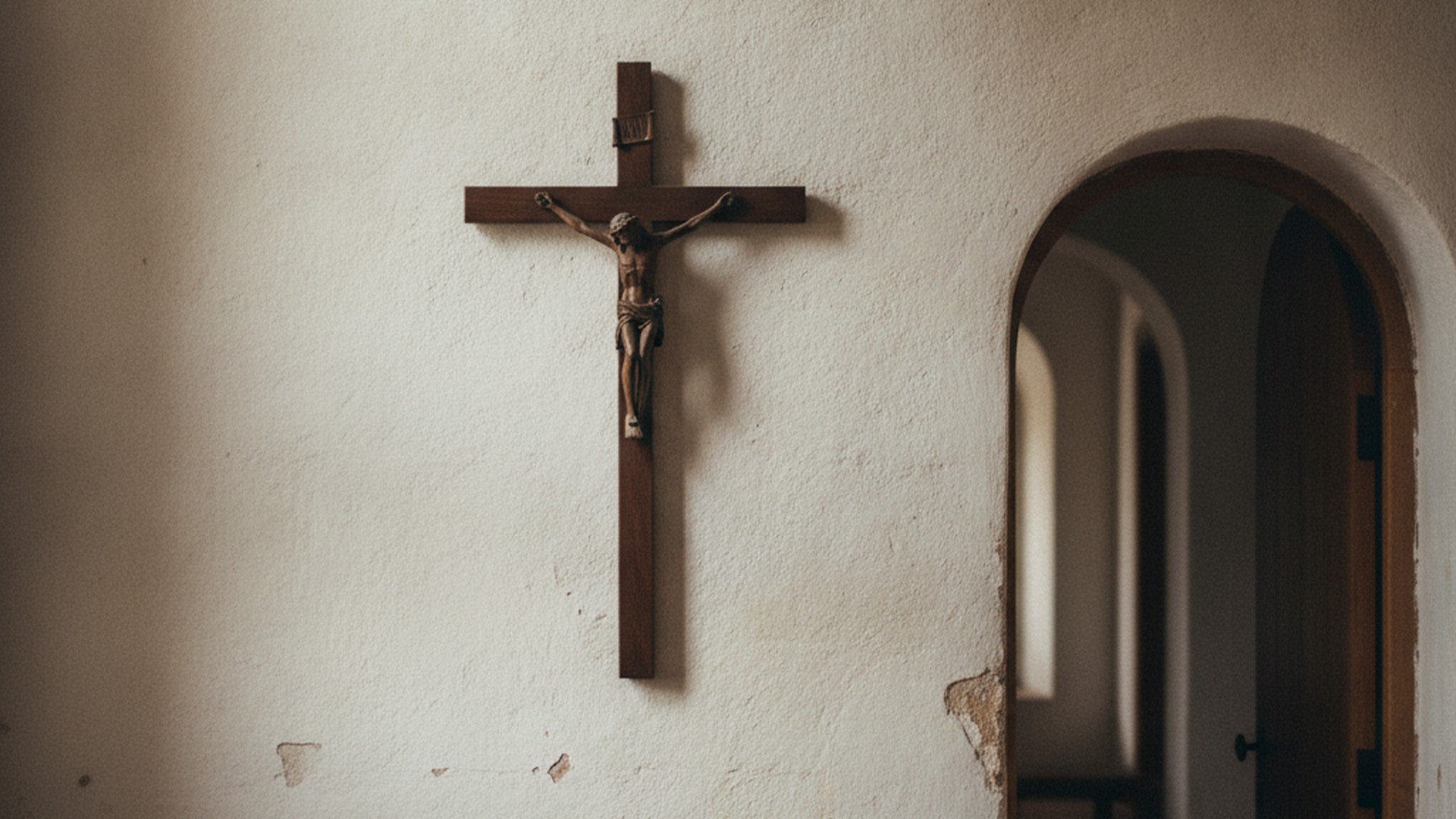 Crucifix de bois sombre accroché à un mur intérieur clair, près d'une embrasure de porte