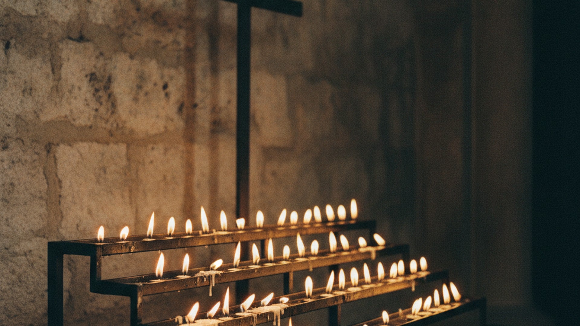 Présentoir de cierges votifs allumés dans la chapelle latérale d'une église de pierre, croix de fer en arrière-plan