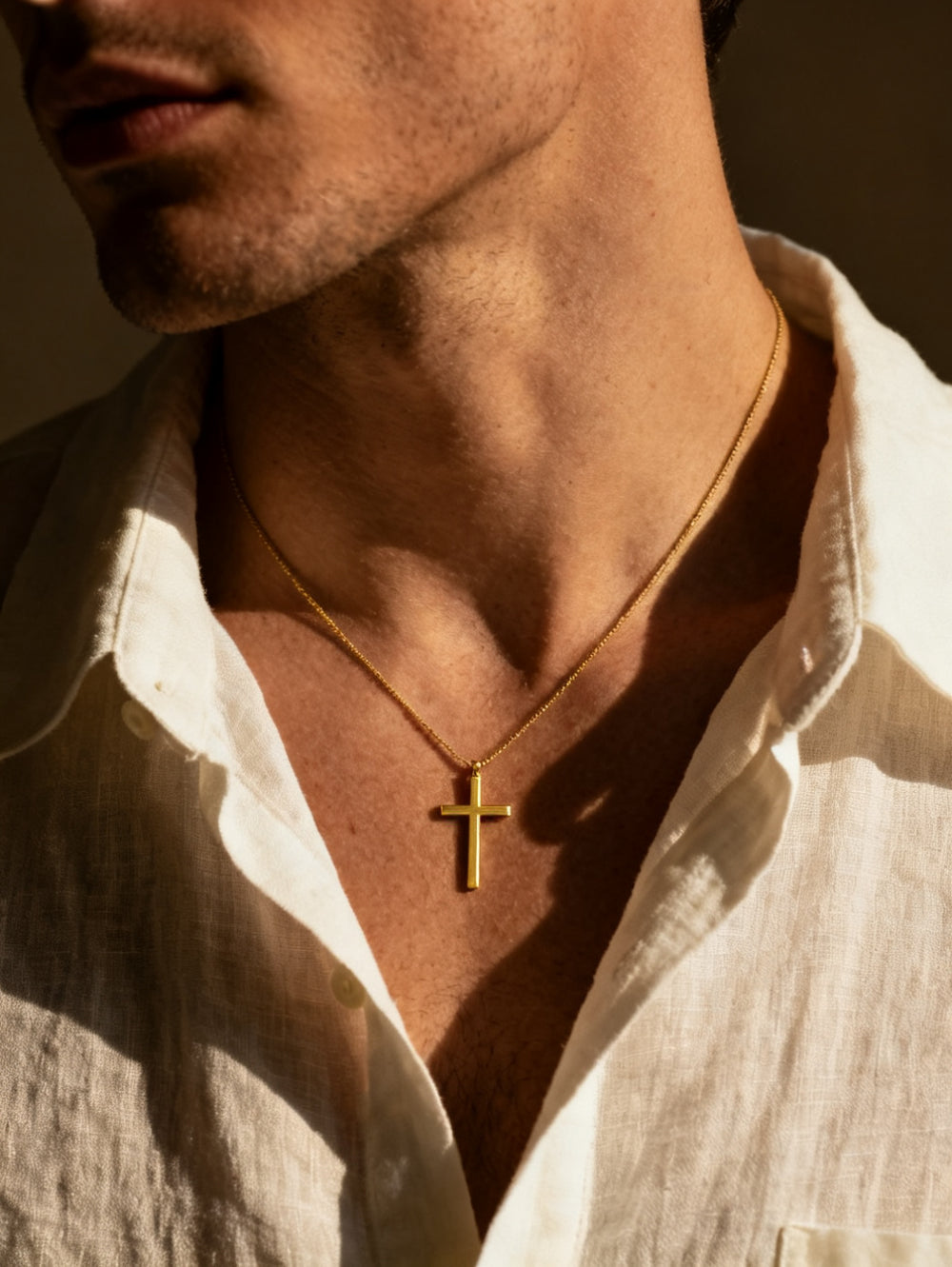 Une fine croix dorée portée sur la peau, chemise de lin, lumière chaude