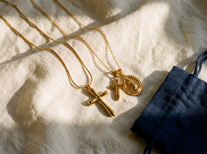 Deux colliers croix et une médaille miraculeuse en doré, posés sur du lin près d'un pochon marine.